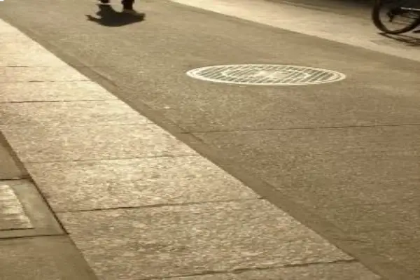 A shadow of a person walking on a street with a manhole cover and a bicycle wheel visible.
