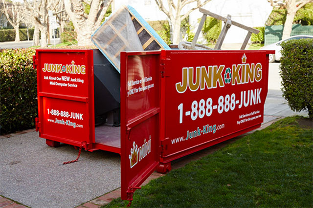 Junk King Dupster on a lawn/sidewalk with a board object leaning up inside