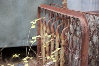 Old rusty bedspring frame leaning against a wall with weeds growing nearby.