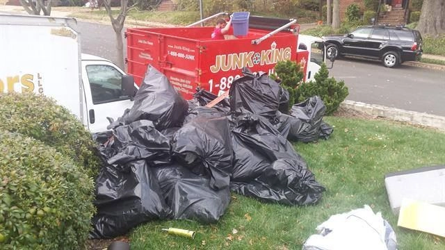 Pile Of Black Trash Bags On A Lawn Next To A Red Dumpster And A White Truck.